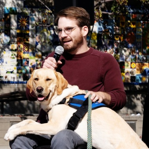 Luke speaking into a microphone on a stage. His lab Glynda is in his lap. Luke is in a manual wheelchair.